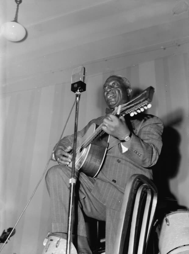 Lead Belly playing his twelve string guitar and singing into a microphone at the National Press Club